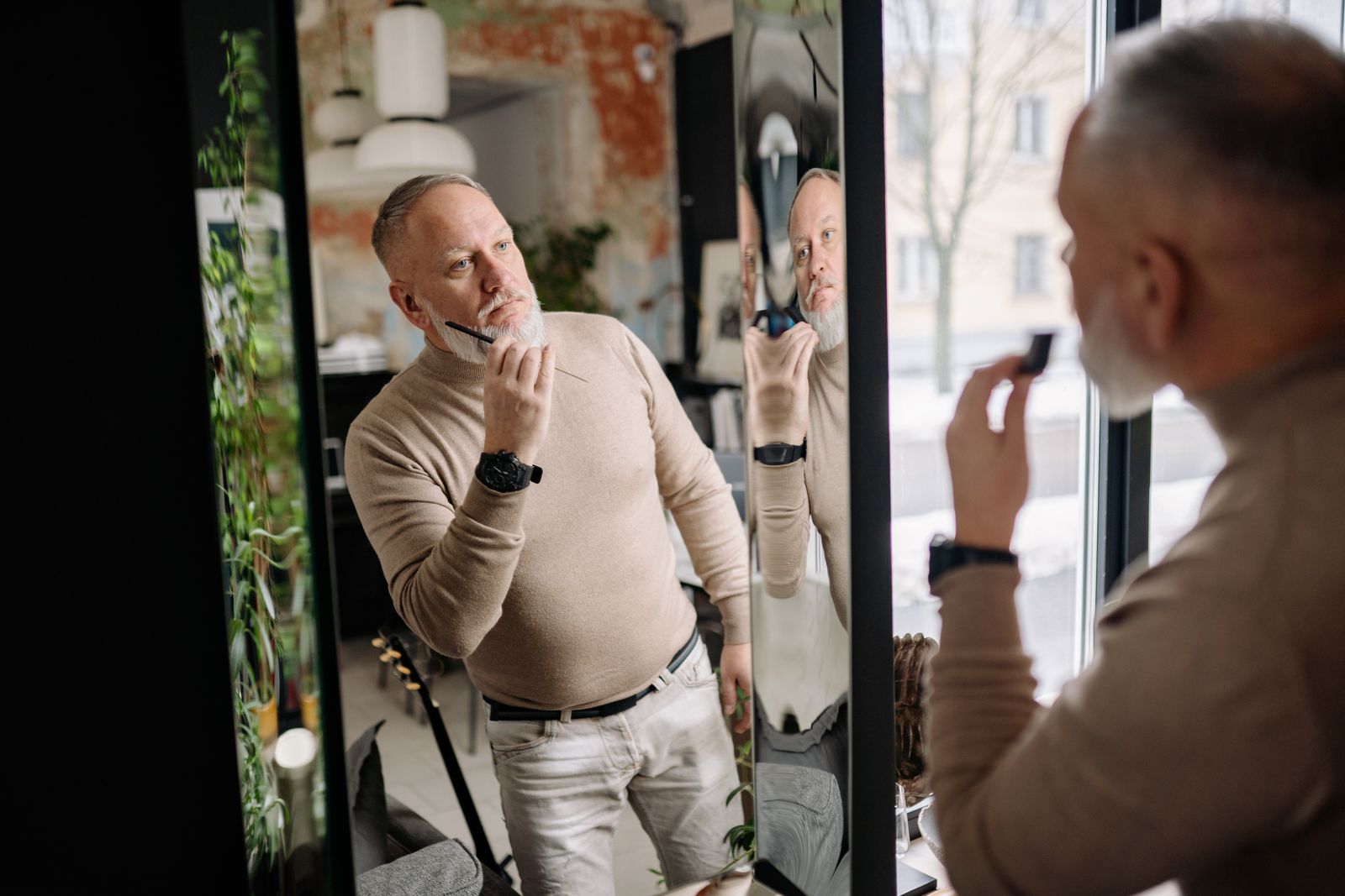 Man i beige tröja som borstar skägget framför en spegel