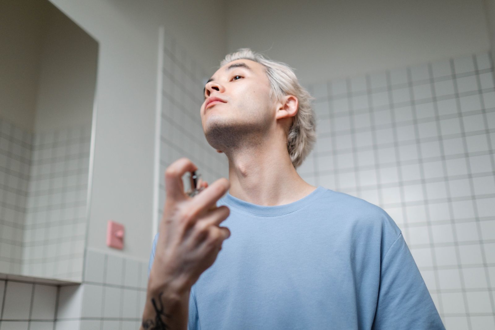 Man med blekt hår och ljusblå t-shirt som sprayar parfym på halsen i ett kaklat badrum